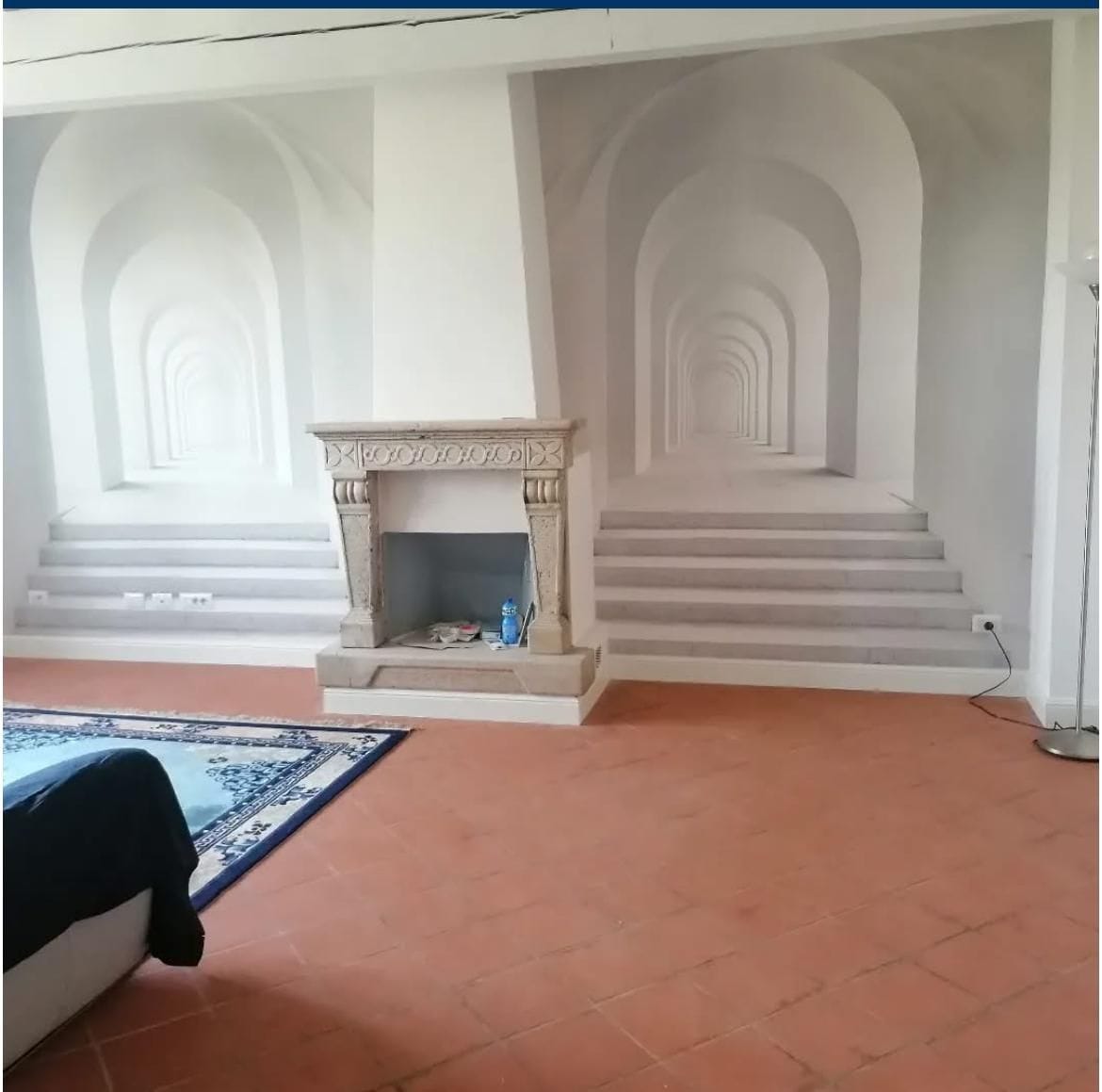 Parete con carta da parati prospettica ai lati di un camino in pietra, pavimento in cotto